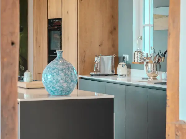 Cuisine équipée aux façades bleu profond et plan de travail clair, sous poutres apparentes.