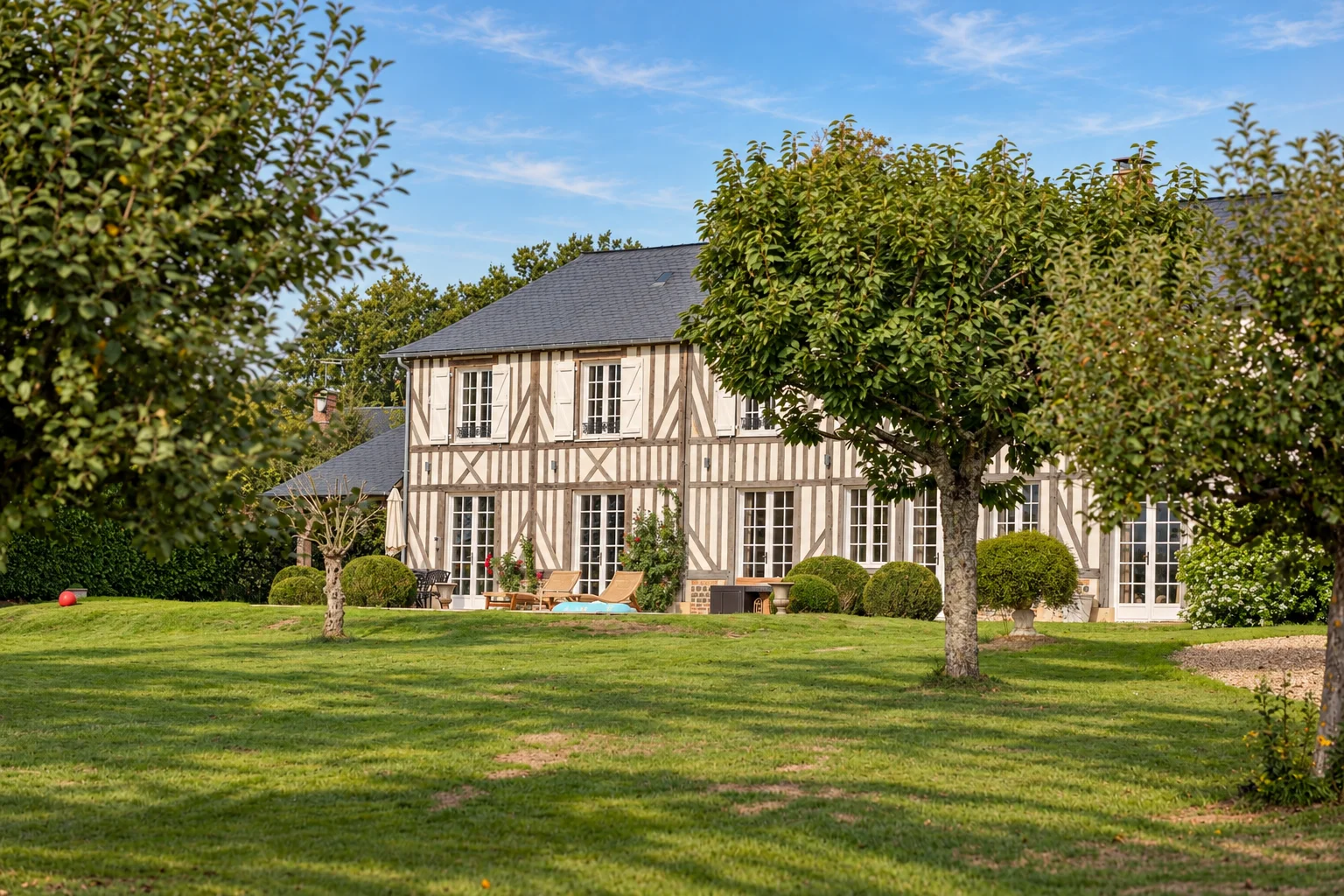 Vue de la maison principale à colombages depuis la pelouse, entre les arbres du jardin.